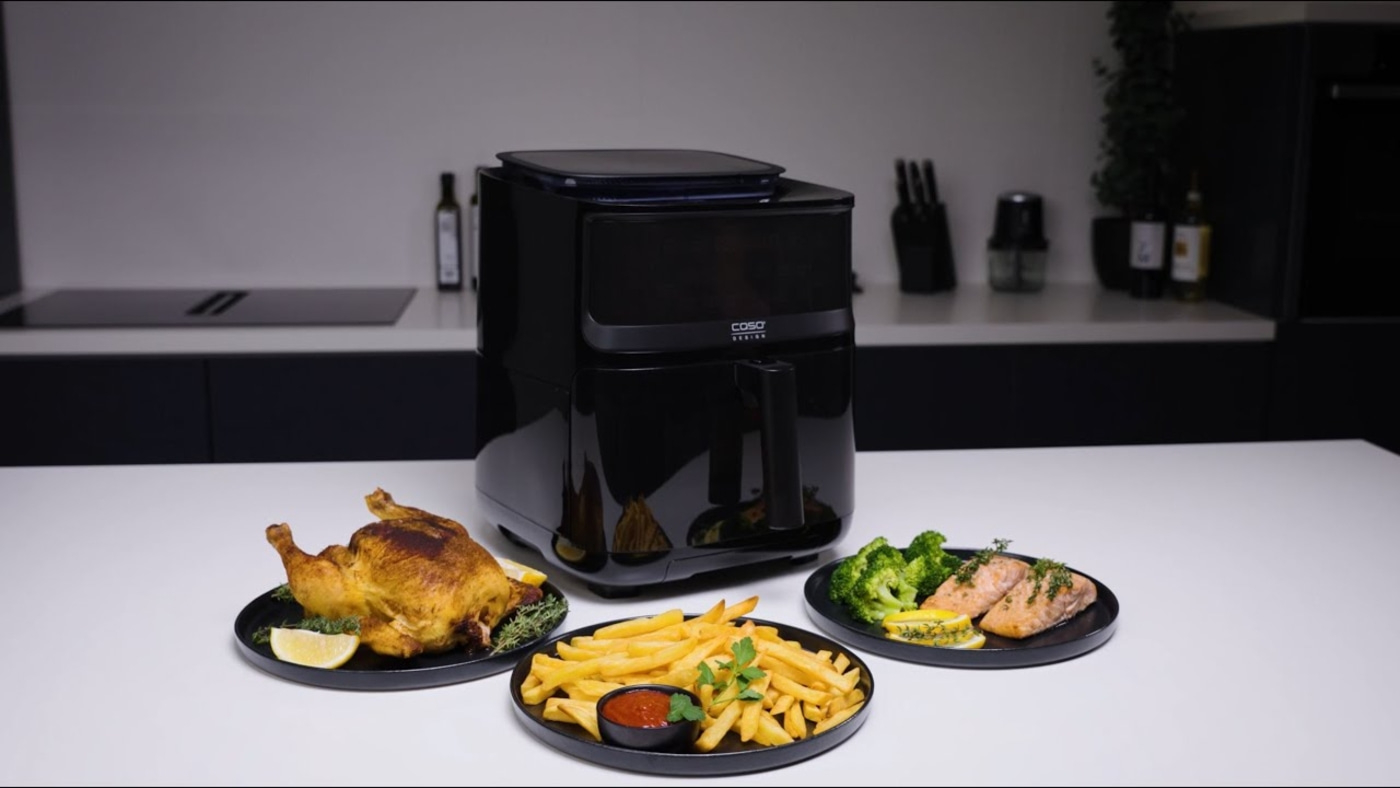 Black air fryer with roasted chicken, fries, and salmon on a table in a kitchen.