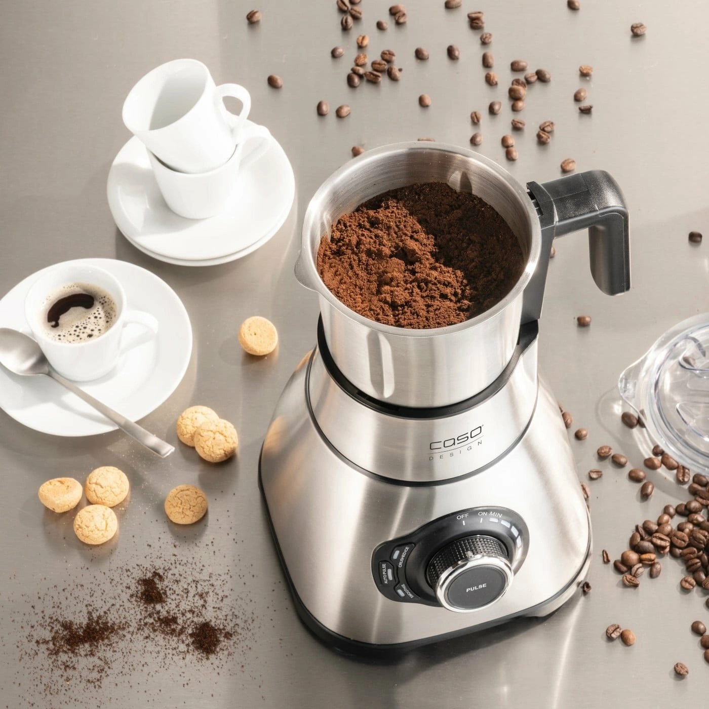 Kaffeemühle mit Kaffee, Tassen, Keksen und Kaffeebohnen auf einer Oberfläche.